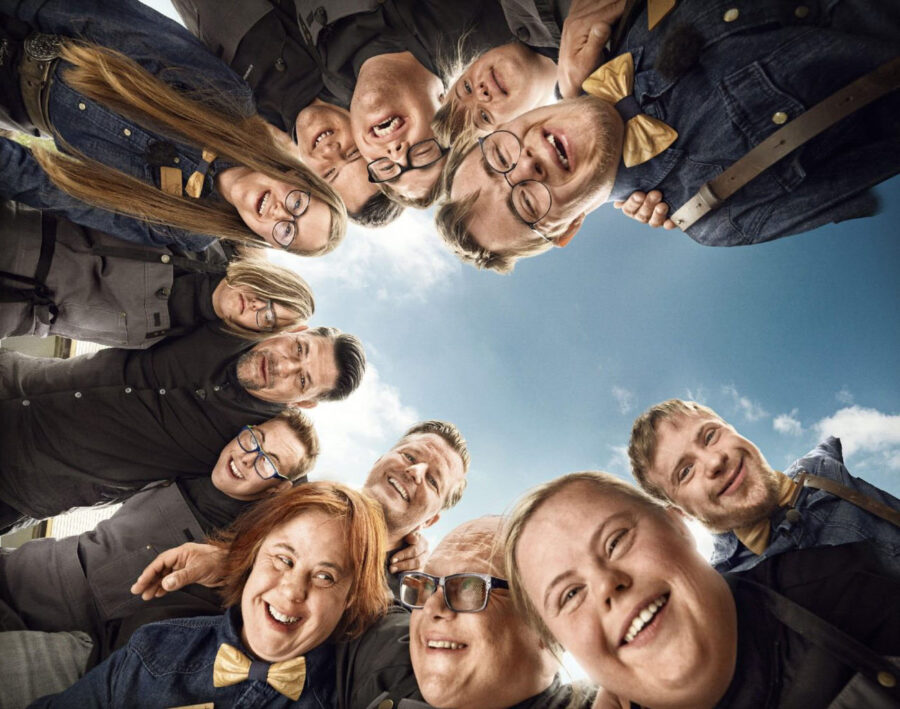 Filmplakat "Zum Schwarzwälder Hirsch" 13 personen stehen im Kreis und blicken nach unten die Kamera, blauer Himmel über ihnen.