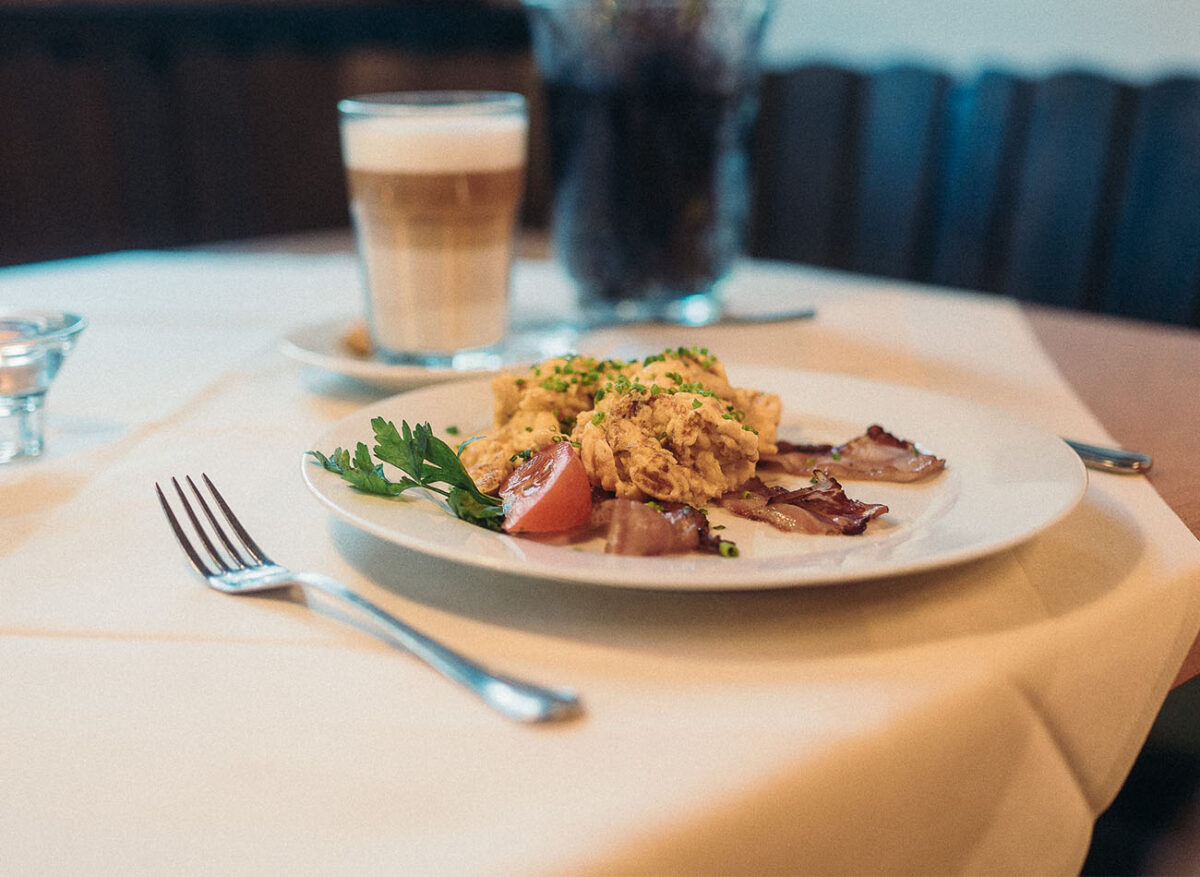 Frühstückteller mit Rührei und Schinken. Im Hinetrgrund steht eine Latte Macchiato