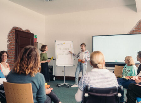 Workshop. Zuhörer:innen sitzen im Stuhlkreis. Zwei Referentinnen am Flipchart erklären Methoden im Online-Raum