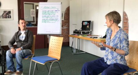 Stuhlkreis im Schulungsraum. Die Referentin für Stimmtraining demonstriert einem Teilnehmenden, der neben ihr sitzt, eine aufrechte Sitzhaltung.