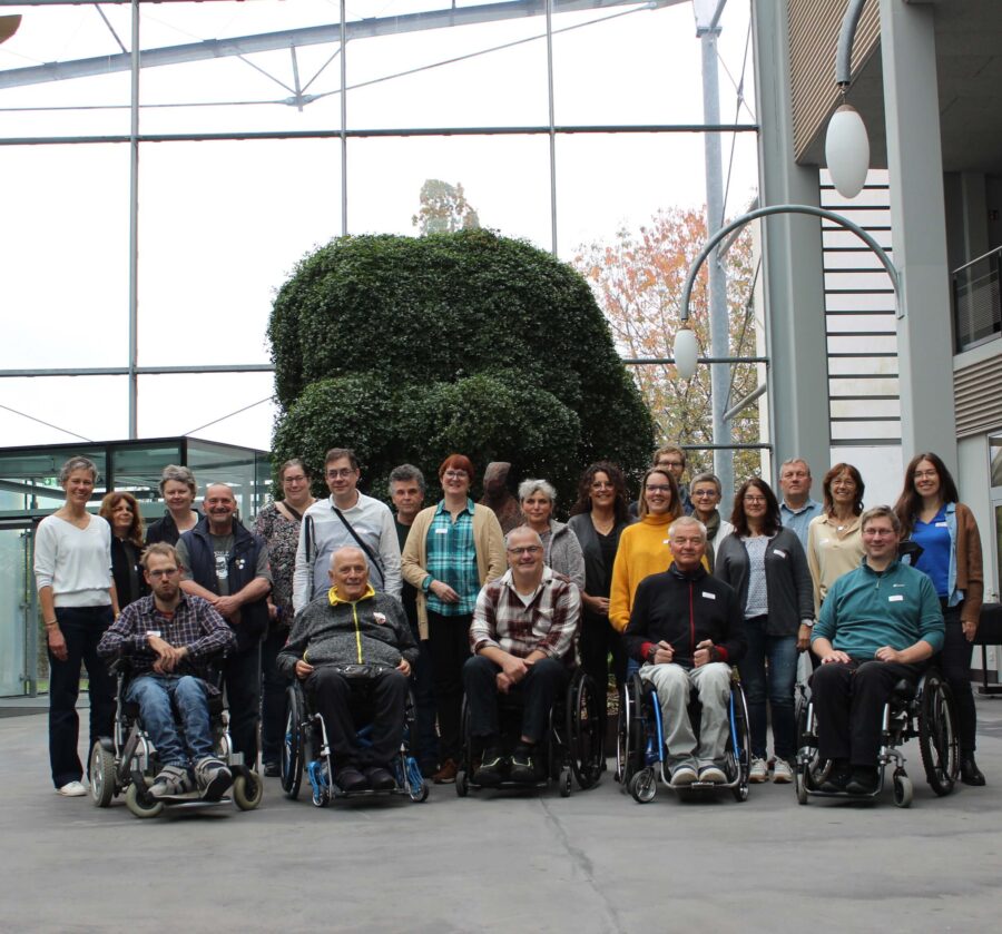 Gruppenbild mit 22 Frauen und Männern in der verglasten Eingangshalle des Tagungshauses, in dem die Basisqualifizierung der neuen KIV stattfindet. In der ersten Reihe fünf Personen mit Rollstuhl.