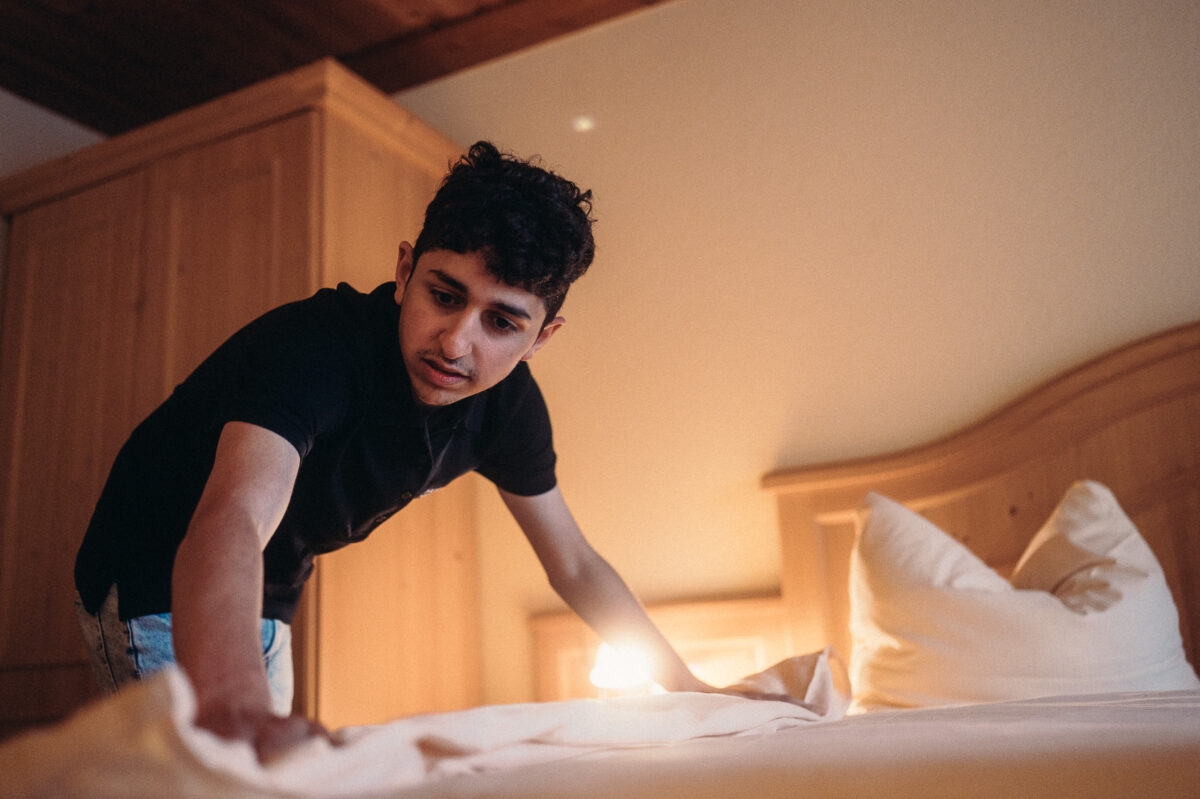 Junger Mann bezieht ein Bett. Im Hintergrund erkennt man ein Kissen und ein Kleiderschrank